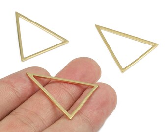 Puede incluir: Tres colgantes triangulares dorados con marco abierto. Uno se sostiene en una mano, mientras que otros dos están dispersos cerca. Las formas geométricas son probablemente para la fabricación de joyas o manualidades.