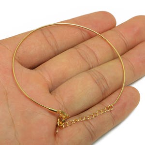 May include: A gold-colored, thin wire bracelet with a delicate chain clasp. The bracelet is held in a hand, showcasing its simple, elegant design. The jewelry piece is likely for everyday wear.
