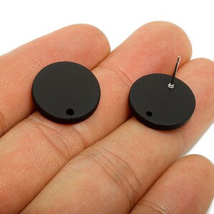 May include: Two matte black, circular stud earrings. One earring has a post for insertion, while the other is a blank disc. Each earring has a small hole near the edge. The earrings are shown against a neutral background.