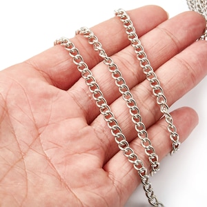 May include: Silver curb chain, a type of chain with flat, interlocking links, shown in a hand. The chain is approximately 6mm wide.