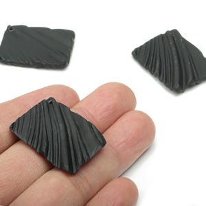 May include: Three black, square pendants with a textured, ridged surface. One pendant is held in a hand, while two others are placed on a white background. Each pendant has a small hole at the top.