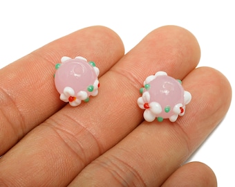 May include: Two pink glass beads with white, red, and green floral accents. The beads are round and appear to be handmade. The beads are displayed on a person's hand.