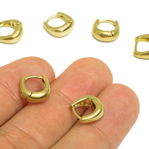 Puede incluir: Se muestran varios pendientes de aro pequeños de color dorado. Dos pendientes se sostienen en una mano, mientras que otros cuatro están dispersos sobre un fondo blanco. Los pendientes tienen forma cuadrada redondeada.