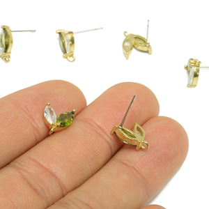 May include: Gold-tone stud earrings with a leaf design. Each earring features two marquise-cut gemstones, one white and one green. The earrings are shown on a white background.