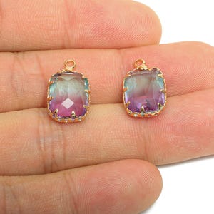 May include: Two gold-toned, rectangular pendants with a faceted, color-shifting gemstone. The gems transition from purple to teal. Each pendant has a small loop at the top for attaching to a chain or other jewelry component.
