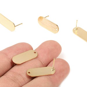 Puede incluir: Postes de pendientes dorados, de forma ovalada, con un poste y un pequeño agujero para fijar componentes de joyería. La imagen muestra varios postes, algunos con una superficie texturizada, y dos sostenidos en una mano. Estos son suministros para la fabricación de joyas.