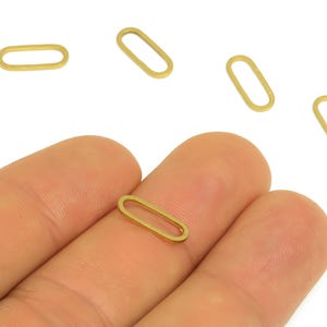 May include: Several small, gold-colored oval-shaped jewelry findings are displayed against a white background. One finding is held in a person's hand. The findings are open ovals, suitable for jewelry making.