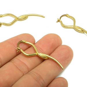 May include: Gold-colored hoop earrings with a unique, flowing design. The earrings have a curved shape with a knot-like detail in the center and a slender, elongated end. The earrings are shown against a white background.