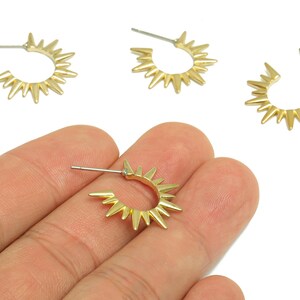 May include: Gold-colored hoop earrings with a sunburst design. The earrings have a post backing and a series of pointed spikes around the outer edge. Several earrings are visible, with one held in a hand.