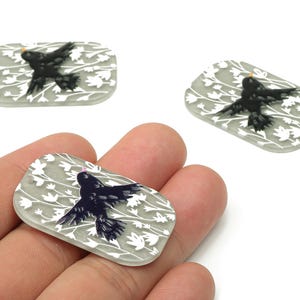 May include: Three rectangular, gray-toned pendants with a black bird design. The bird is in flight, set against a white floral pattern. One pendant is held in a hand. The pendants are likely for jewelry making.