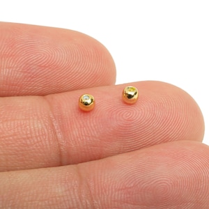 May include: Two small, gold-colored beads with a small, clear gem set in each. The beads are round and appear to be made of metal. They are resting on a person's fingertip.