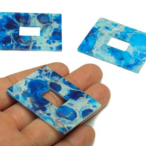 Puede incluir: Tres colgantes rectangulares de resina veteada azul y blanca con un recorte rectangular. Un colgante se sostiene en una mano. Los colgantes tienen un patrón veteado de remolinos azules y blancos.
