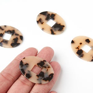 Peut inclure: Perles ovales avec un motif beige clair et marron foncé, avec une découpe carrée au centre. Une perle est tenue dans une main, avec trois autres dispersées sur une surface blanche. Les perles ont un motif écaille de tortue.