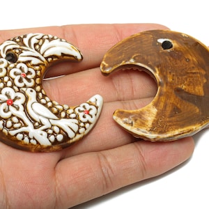 May include: Two ceramic crescent moon pendants. One is brown with white floral and bird details, and the other is a solid brown colour. Both have a hole for stringing.