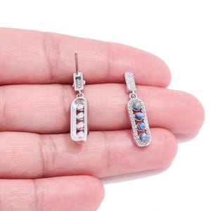 Puede incluir: Un par de pendientes colgantes plateados. Uno tiene tres piedras claras, el otro tres piedras opalescentes. Ambos tienen un diseño rectangular con una pequeña parte superior decorativa.