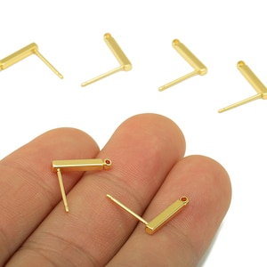 May include: Gold-colored, L-shaped earring posts with a rectangular bar and a post for insertion. Several pieces are scattered on a white background, with two held in a hand. The earrings are small and minimalist.