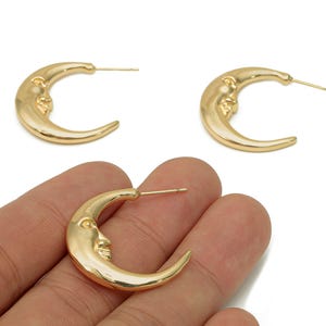 May include: Gold-colored crescent moon earrings with a detailed face design. The earrings are in a hoop style and have a post backing. The image shows three earrings, with one held in a hand. The earrings are a celestial-themed jewelry item.