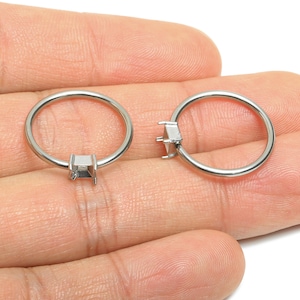 May include: Two silver-colored ring blanks with square settings are displayed. The rings are held in a hand, showcasing their simple, modern design. These rings are ideal for jewelry making and crafting.