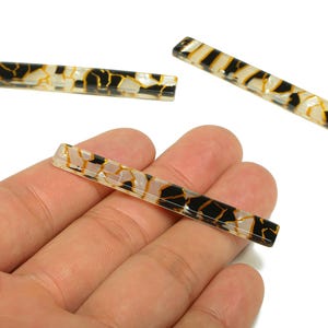 Könnte beinhalten: Rechteckige dekorative Perlen mit einem schwarzen, goldenen und weißen Muster. Die Perlen werden in einer Hand gezeigt und sind etwa 2,5 cm lang. Die Perlen haben eine glänzende Oberfläche und eignen sich für Schmuckherstellung oder Bastelprojekte.