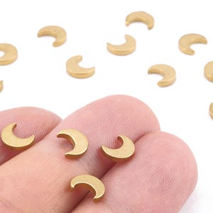 May include: Small, gold-colored crescent moon charms. Several charms are scattered on a white surface, with a few held in a person's fingers. These charms are likely used for jewelry making or other craft projects.