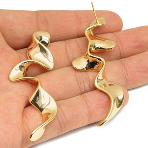 May include: A pair of gold-coloured, abstract earrings with a flowing, ribbon-like design. The earrings have a post backing and a polished, reflective surface. The jewellery is displayed against a neutral background.