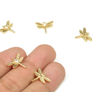 Op de afbeelding: Gouden libelle bedels met een heldere kristallen accent. De bedels zijn klein en gedetailleerd, met delicate vleugels en een slank lichaam. Er zijn verschillende bedels te zien, sommige worden in een hand gehouden, tegen een witte achtergrond.