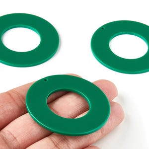 Op de afbeelding: Drie groene acryl cirkelhangers met een gat voor het maken van sieraden. De hangers hebben een effen, glanzende groene kleur. Eén hanger wordt in de hand gehouden, terwijl de andere twee op een wit oppervlak liggen.