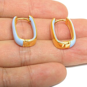 May include: A pair of gold-colored hoop earrings with a rectangular shape. The earrings have a light blue enamel filling and a hinged closure. The earrings are shown on a person's hand.