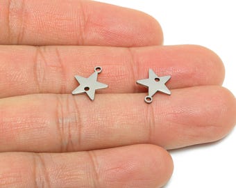 Puede incluir: Dos pequeños dijes de estrella de color plateado. Cada dije tiene forma de estrella de cinco puntas con un pequeño agujero en el centro y un lazo en la parte superior para sujetarlo a la joyería. Los dijes se muestran sobre un fondo color piel.