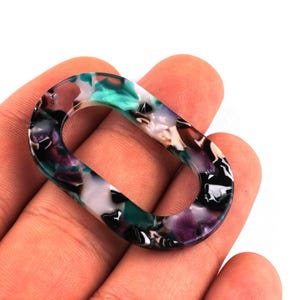 Peut inclure: Un maillon ovale en acrylique dans les tons de turquoise, de violet, de noir et de blanc. Le motif marbré est visible sur tout le maillon. Le maillon est tenu dans la main d'une personne sur fond blanc. Cet article convient à la fabrication de bijoux.