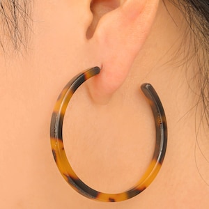 May include: A close-up of a tortoise shell hoop earring. The earring is a large, open-ended circle with a brown and yellow pattern. The earring is worn on a person's ear, with the ear and hair visible in the background.