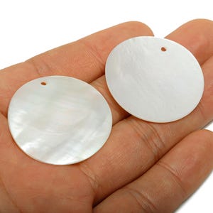Peut inclure: Deux pendentifs ronds en nacre blanche avec un trou en haut sont tenus dans une main. Les pendentifs ont un éclat nacré et une surface lisse. Ces pendentifs sont idéaux pour la fabrication de bijoux.