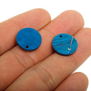 May include: Two round, blue earrings with a marbled effect are displayed. One earring has a post attached, while the other has a small hole. The earrings are held in a person's hand.