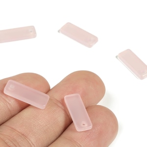 Puede incluir: Se muestran varias cuentas rectangulares de color rosa pálido con un agujero en un extremo. Las cuentas son lisas y translúcidas, con un color suave y uniforme. Algunas cuentas se sostienen en una mano, otras están dispersas.