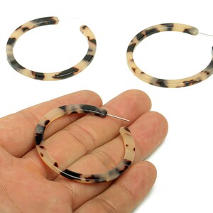 May include: Three hoop earrings with a tortoiseshell pattern. The earrings are a mix of cream, black, and brown. One earring is held in a hand, while the other two are displayed on a white background. The earrings have a silver post.