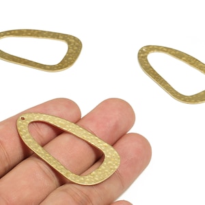 Puede incluir: Tres colgantes dorados de marco abierto con textura martillada. Un colgante se sostiene en una mano, mientras que los otros dos están colocados cerca. Los colgantes tienen una forma ovalada irregular con un pequeño agujero en la parte superior.