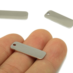 Puede incluir: Etiquetas rectangulares de metal plateado con un pequeño agujero en un extremo. Se ven tres etiquetas, una sostenida en una mano. Las etiquetas tienen esquinas redondeadas y una superficie lisa y reflectante. Ideales para la fabricación de joyas o manualidades.