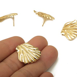 Puede incluir: Pendientes de botón dorados con forma de concha. Los pendientes tienen una superficie detallada y texturizada y un poste para orejas perforadas. La imagen muestra los pendientes sobre un fondo blanco, con un pendiente en la mano.