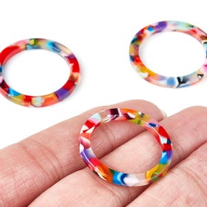 May include: Three colorful resin rings with a marbled pattern in red, blue, yellow, and purple hues. One ring is held in a hand, while two others are displayed on a white background. These rings are suitable for jewelry making.