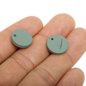 May include: Two sage green, round stud earrings. One earring has a post, the other has a small hole. The earrings are displayed on a hand against a white background. Jewellery.