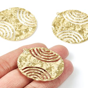 Könnte beinhalten: Drei goldfarbene, strukturierte Metallanhänger mit runder Form und einem ausgeschnittenen Design aus gebogenen Linien. Die Anhänger haben oben eine kleine Schlaufe zum Befestigen an einer Kette oder einem anderen Schmuckbestandteil. Die Anhänger werden auf weißem Hintergrund gezeigt.
