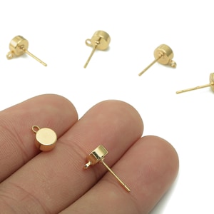 Könnte beinhalten: Goldfarbene Ohrstecker mit flachem, rundem Oberteil und einem Stift zum Einsetzen. Mehrere sind auf weißem Hintergrund verstreut, einige werden in einer Hand gehalten. Die Ohrringe haben eine kleine Schlaufe auf der Rückseite des runden Oberteils.