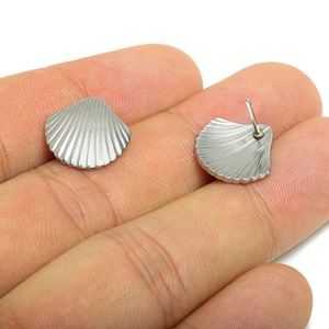 May include: A pair of silver-colored seashell stud earrings. Each earring is shaped like a scallop shell with detailed ridged lines. The earrings are displayed on a hand against a white background.