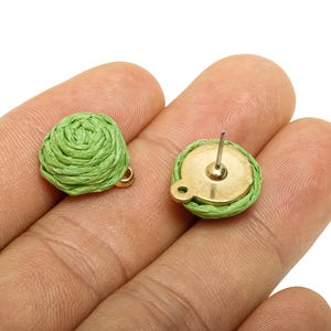 May include: A pair of light green woven stud earrings. The earrings have a round shape with a woven texture and a gold-coloured backing. One earring is face up, and the other is face down, showing the post.