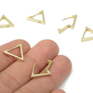 May include: Gold-colored triangle hoop earrings. The earrings are geometric in shape, with a hinged closure. Several earrings are displayed, some open and some closed, against a white background.