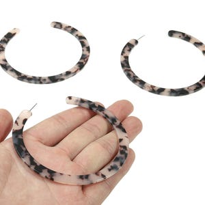 May include: Three pairs of large hoop earrings with a tortoiseshell pattern. The earrings are a mix of black, brown, and cream colors. One pair is held in a hand, while the other two pairs are displayed on a white background.