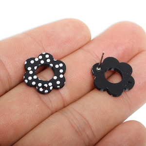 May include: A pair of black flower-shaped stud earrings. One earring has a white polka dot pattern, the other shows the back with a silver post. The earrings are held in a hand.
