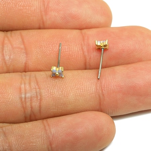 May include: Two small stud earrings with gold-colored square settings and silver posts. The settings feature a light blue-gray stone. The earrings are displayed on a person's hand.