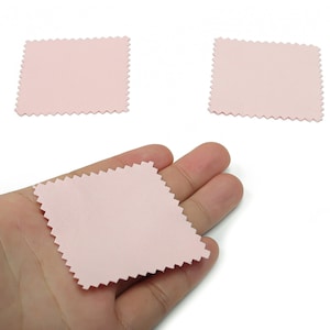 May include: Three light pink, square-shaped cleaning cloths with a zig-zag edge. One cloth is held in a hand, while two others are positioned above. The cloths are on a white background.