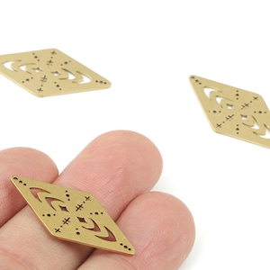 May include: Three gold-coloured diamond-shaped metal charms with decorative cut-out designs. One charm is held between fingers, while two others are positioned in the background. The charms feature crescent and dot patterns.